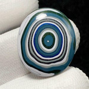 May include: A round, blue, green and white agate cabochon with a concentric circle pattern. The cabochon is polished and has a glossy finish.