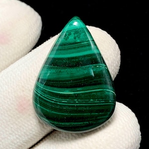 Puede incluir: Una piedra preciosa de malaquita pulida en forma de lágrima. La piedra presenta bandas de color verde oscuro y claro, creando un patrón rayado. La gema se sostiene sobre un fondo de tela blanca.