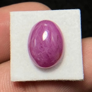 Può includere: Una gemma di rubino lucidata, di forma ovale, che mostra una tonalità rosso-rosata intensa. La gemma è posta su un quadrato bianco, con inclusioni interne visibili e una superficie riflettente. La gemma misura circa 2,5 cm di lunghezza.