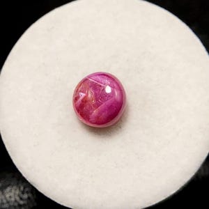 Op de afbeelding: Een ronde, gepolijste robijn edelsteen, met een diepe roze-rode tint met interne lichtreflecties. De edelsteen is geplaatst tegen een witte, getextureerde achtergrond, die de levendige kleur en helderheid benadrukt. Ideaal voor het maken van sieraden.