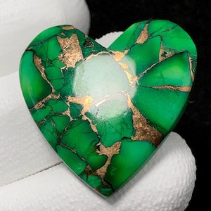 May include: A heart-shaped green turquoise cabochon with gold inclusions. The stone has a polished finish and is set in a heart-shaped setting.