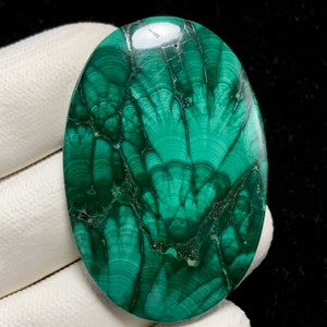 Könnte beinhalten: Ein grüner Malachit-Cabochon mit einem einzigartigen, wirbelnden Muster. Der Stein ist oval geformt und hat eine polierte Oberfläche.
