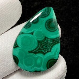 Peut inclure: Une pierre précieuse de malachite polie en forme de goutte d'eau. La pierre présente une couleur verte vibrante avec des motifs complexes et tourbillonnants de vert plus foncé et de noir. Le bijou est tenu par une main gantée de blanc.