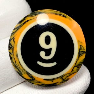 May include: A round, yellow, black, and white billiard ball with the number 9 in the center. The ball has a black center with a white number 9. The outer ring is yellow with black marbling.