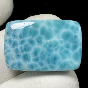 May include: A rectangular, polished gemstone with a light blue hue and a unique, mottled pattern. The stone's surface has a glossy finish, and the color is reminiscent of ocean water. The gemstone is set against a white background.