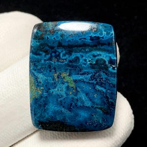 Peut inclure: Une pierre précieuse rectangulaire polie, d'un bleu vibrant, ornée de motifs bleus foncés, noirs et verts. La surface de la pierre est lisse et réfléchissante, révélant des formations minérales naturelles. La pierre est tenue sur un fond blanc.