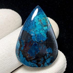 Puede incluir: Una piedra preciosa pulida en forma de lágrima con una llamativa mezcla de colores. La piedra presenta una base negra profunda y brillante, superpuesta con vibrantes patrones turquesa y verde azulado. La gema se sostiene sobre un fondo blanco.