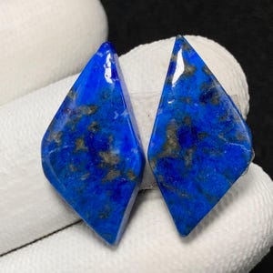 Può includere: Un paio di cabochon di lapislazzuli blu tagliati a forma di diamante. Le pietre hanno un colore blu scuro con macchie bianche e dorate.
