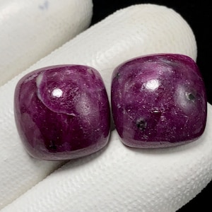 Puede incluir: Dos cabujones de piedra preciosa púrpura con un acabado liso y pulido. Los cabujones son de forma cuadrada y tienen una parte superior ligeramente redondeada.