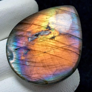 May include: A teardrop-shaped labradorite gemstone with a polished surface. The stone displays a vibrant play of color, with hues of orange, purple, blue, and yellow. The gemstone is held by a gloved hand, showcasing its iridescent qualities.