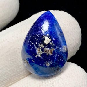 Puede incluir: Una piedra preciosa de lapislázuli en forma de pera con un color azul real profundo y visibles inclusiones metálicas de pirita dorada dispersas por la superficie. La piedra tiene un acabado liso y pulido con una silueta redondeada.