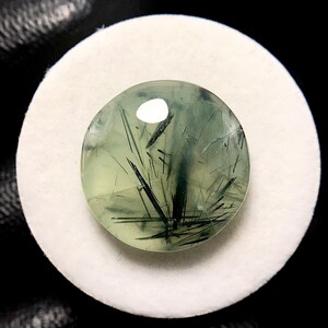 May include: A round, polished gemstone with a pale green hue and dark, needle-like inclusions. The gemstone is set against a white, circular backdrop, with a dark background. The stone's surface reflects light, creating a bright spot.
