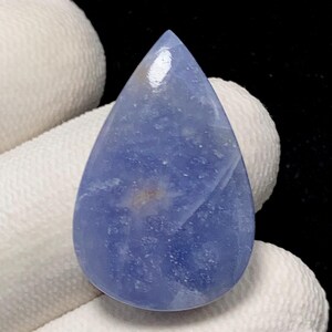 May include: A teardrop-shaped, polished gemstone in shades of blue with subtle marbling and a glossy finish. The stone has a smooth surface and is held against a white background.