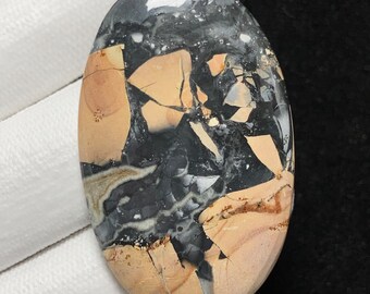 May include: An oval gemstone with a distinctive pattern. The stone displays a blend of black, tan, and cream hues, with a cracked, mosaic-like design. The polished surface reflects light, emphasizing the natural variations within the stone.