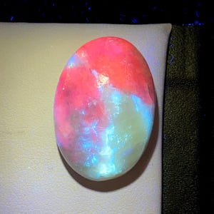 May include: An oval-shaped cabochon gemstone displaying a vibrant play-of-color with intense flashes of neon pink, bright red, and electric blue against a translucent, milky white base. The polished surface reflects light, highlighting the internal structure.