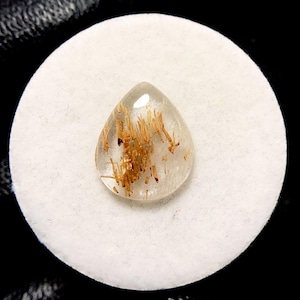 Może przedstawiać: Przezroczysty kamień szlachetny w kształcie łzy ze złotymi, igiełkowatymi inkluzjami. Kamień szlachetny jest osadzony na białym, okrągłym tle, z ciemnym tłem. Inkluzje tworzą uderzającą wizualną teksturę.