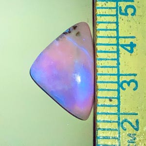 May include: A triangular, polished gemstone with a gradient of pink, purple, and blue hues. The stone is next to a ruler, which measures the stone's length. The gemstone has a smooth surface and a slightly irregular shape.
