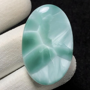 Op de afbeelding: Een ovale cabochon edelsteen met een draaiende patroon in tinten van groen en wit. De steen is gepolijst en heeft een glad oppervlak.