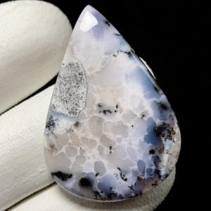 Puede incluir: Una gema pulida en forma de lágrima. La piedra presenta una mezcla de tonos blancos, grises y azules, con patrones similares a nubes e inclusiones dendríticas. Salpicaduras negras salpican la superficie, agregando contraste.