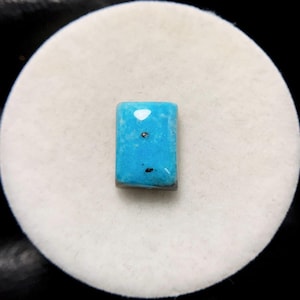 Puede incluir: Una piedra preciosa rectangular de turquesa con una superficie pulida. La piedra tiene un tono azul vibrante con inclusiones blancas y negras. La piedra preciosa está sobre un fondo blanco.