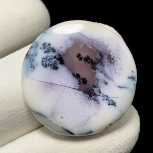May include: A round, polished gemstone with a white base, featuring intricate patterns of lavender and black dendritic inclusions. The stone has a smooth, glossy surface and a natural, organic appearance. The gemstone is held by a white surface.