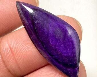 May include: A polished, elongated gemstone with a deep purple hue. The stone has a smooth, glossy surface with subtle variations in color and texture. The shape is tapered, with a rounded end and a pointed tip. The gemstone is held between fingers.
