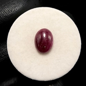 May include: A deep magenta-colored oval gemstone with a smooth, polished cabochon surface. The stone features a translucent quality with subtle internal inclusions and a reflective highlight, centered on a circular white display pad.