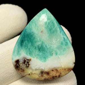 May include: A teardrop-shaped gemstone with a turquoise and white pattern, resembling ocean waves. The bottom edge features a brown and tan band. The stone is held by a white gloved hand, with a black background.