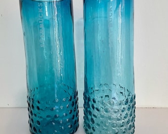 May include: Two teal blue glass tumblers with a textured, bubble-like design. The glasses are empty and appear to be made of recycled glass.