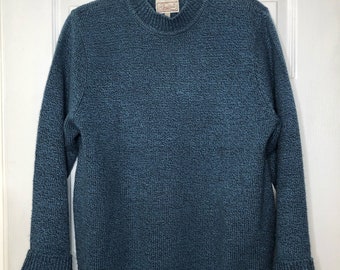 May include: Teal-colored sweater with a textured knit pattern. The sweater has a mock neck, long sleeves, and a small label near the neckline. The sleeves have a flared cuff. The sweater is hanging on a wooden hanger.