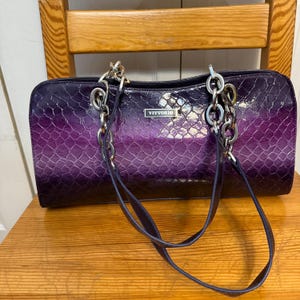 May include: A purple and black handbag with a snakeskin pattern. The bag has silver chain handles and a dark purple strap. The word "Vittorio" is displayed on a silver plaque on the front of the bag. The bag is sitting on a wooden chair.
