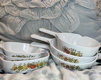 May include: A set of white ceramic cookware with a floral pattern featuring green leaves, red and yellow fruits, and white flowers. The cookware includes a casserole dish, a saucepan, and a frying pan.