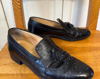 May include: Pair of black leather loafers with tassel accents. The shoes feature a textured vamp and decorative perforations. The loafers are displayed on a wooden surface, showcasing their classic design and craftsmanship.