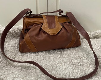 May include: A brown leather satchel bag with a tan leather trim and a long adjustable strap.