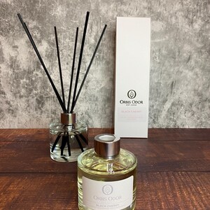 以下が含まれることがあります： 黒い蓋と黒いディフューザーのスティックが付いた透明なガラス瓶。瓶は薄い黄色の液体で満たされており、「Orbis Odor Est. 2020 Black Cherry Aromatic Reed Diffuser」と書かれたラベルが付いています。