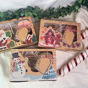 May include: Three gift boxes with Christmas themes. The boxes are decorated with images of Santa Claus, a gingerbread house, and a snowman. Each box has a tag attached to it. The boxes are sitting on a white surface with a candy cane.