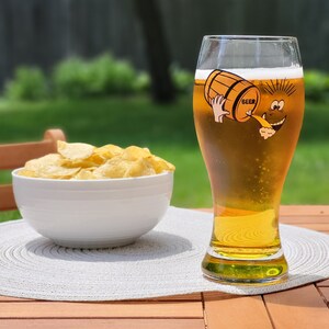 May include: A tall glass of beer with a cartoon image of a barrel pouring beer into a glass. The glass has the word "BEER" on the barrel. A bowl of potato chips is in the foreground.