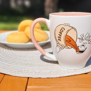 May include: White ceramic coffee mug with a pink handle. The mug features a cartoon illustration of a person with a cup of coffee pouring into their mouth. The text "COFFEE" is printed on the side of the cup.