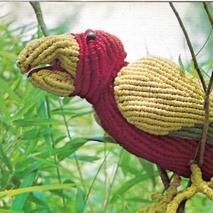 Puede incluir: Escultura de pájaro hecha a mano con ganchillo. El pájaro tiene un cuerpo rojo, pico y patas amarillas, y alas verdes y amarillas. Está posado en una rama delgada en medio de follaje verde, mostrando un diseño vibrante y texturado.