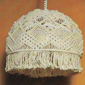 Può includere: Un paralume bianco lavorato a crochet con un bordo a frangia appeso a un cavo bianco. Il paralume ha un motivo geometrico.