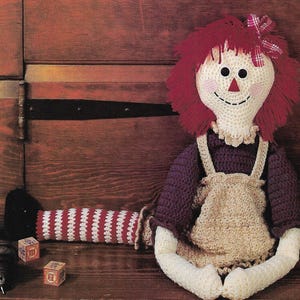 Pode incluir: Uma boneca de pano de crochê com cabelo vermelho, um vestido marrom e pernas listradas de branco e vermelho. A boneca está sentada em uma superfície de madeira.