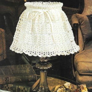 May include: A white crocheted lampshade with a white ribbon tied around the top. The lampshade is sitting on a table with a white crocheted table runner and two white crocheted doilies.