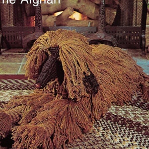 May include: A brown Afghan hound dog sculpture made of yarn, lying on a patterned rug in front of a fireplace. The text "DUKE OF PEDIGREE The Afghan" is visible at the top of the image.