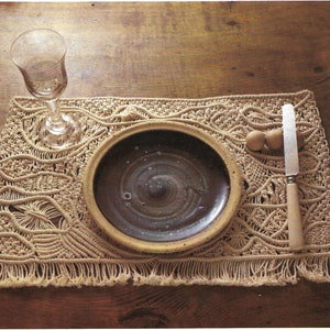 May include: A brown ceramic plate with a dark brown glaze sits on a beige crocheted placemat with fringe. A glass wine glass and a wooden handled knife are also on the placemat.