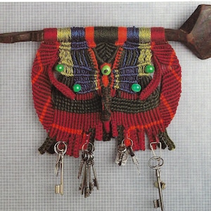 Op de afbeelding: Een kleurrijk macramé wandkleed in de vorm van een vlinder met groene kralen voor ogen. Het kleed is bevestigd aan een houten stok met een houten lepel aan het uiteinde. Een set sleutels hangt aan de onderkant van de vlinder.