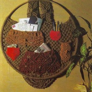 May include: A brown macrame wall hanging with multiple pockets. The pockets are made of different colored macrame and are filled with various items, including letters, pens, and a magazine. The wall hanging is circular and has a wooden hoop at the top.