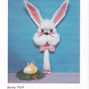 May include: A crocheted bunny wall hanging with pink ears, white fur, and a red and white gingham bow. The bunny is holding a white tassel with pink beads. A yellow and white Easter egg is in the foreground.