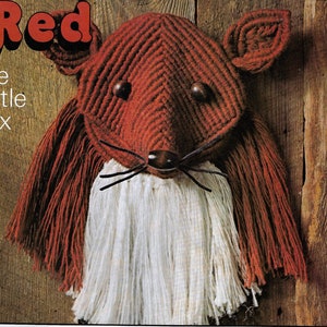 以下が含まれることがあります： 「Red the Little Fox」というタイトルの赤いマクラメの狐が、木の壁に飾られています。狐は赤と白の糸でできており、白いひげと茶色の鼻をしています。