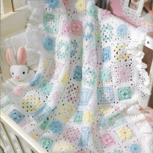 Könnte beinhalten: Eine weiße gehäkelte Babydecke mit einem gekräuselten Rand. Die Decke besteht aus Quadraten in verschiedenen Pastellfarben, darunter Rosa, Blau, Gelb und Grün. Die Decke liegt in einem Kinderbett mit einem weißen Hasenspielzeug.