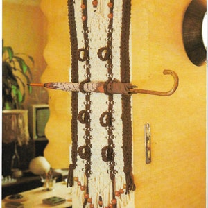 May include: A white and brown macrame wall hanging with wooden beads. The hanging has a fringe at the bottom and is attached to a wall with a wooden dowel. An umbrella is resting on the hanging.